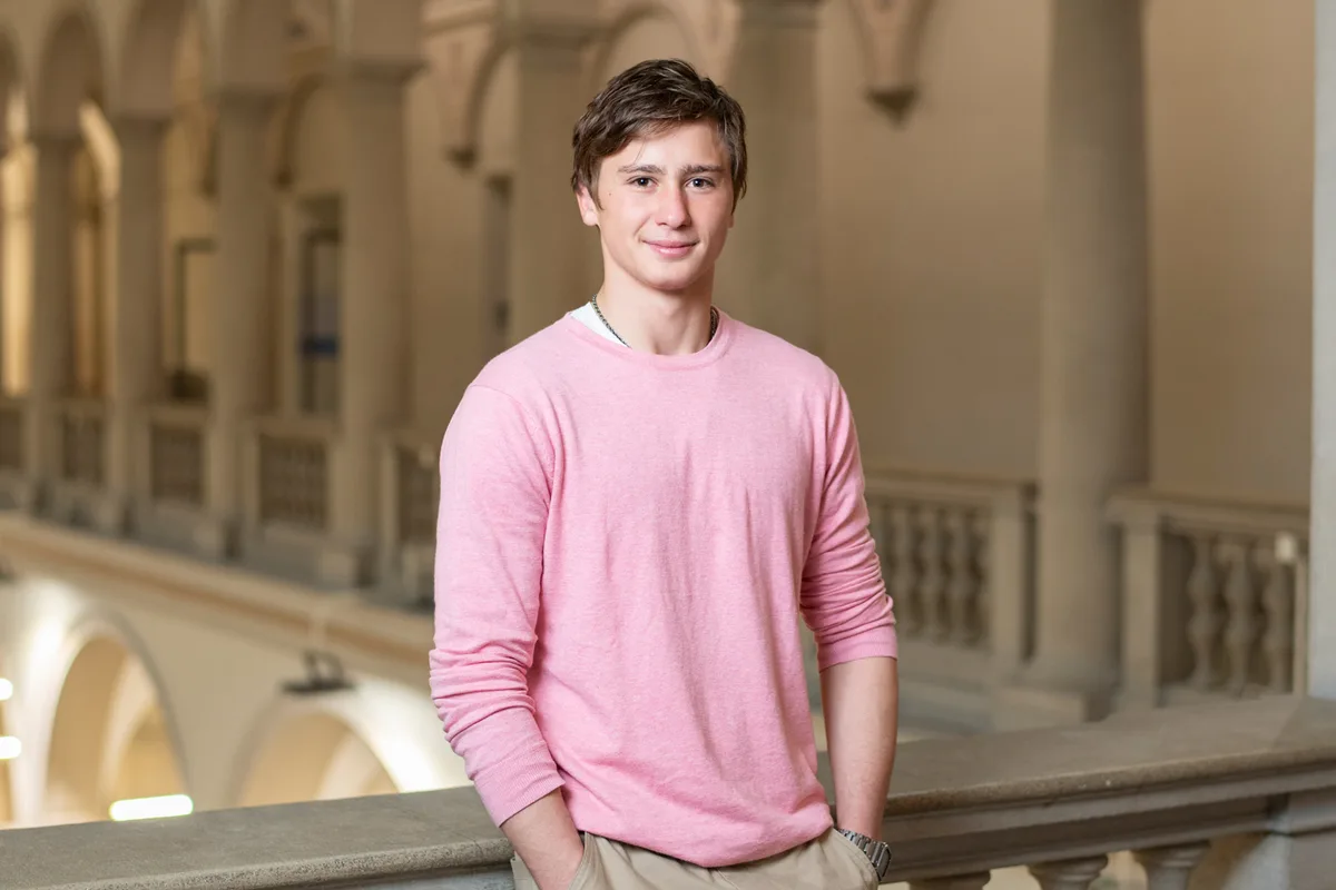 Porträt eines jungen Mannes. Er lehnt an ein Geländer im Innenhof einer Universität.